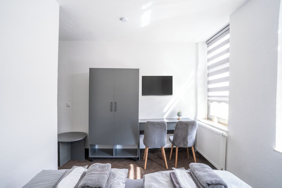 A modern hotel room with a bed, a desk, a chair, and a wardrobe.