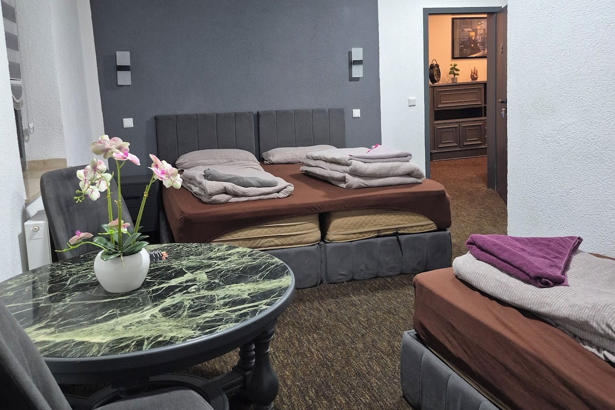 A modern bedroom with a bed, a sofa, a dining table with a vase of flowers, and a small kitchen area.
