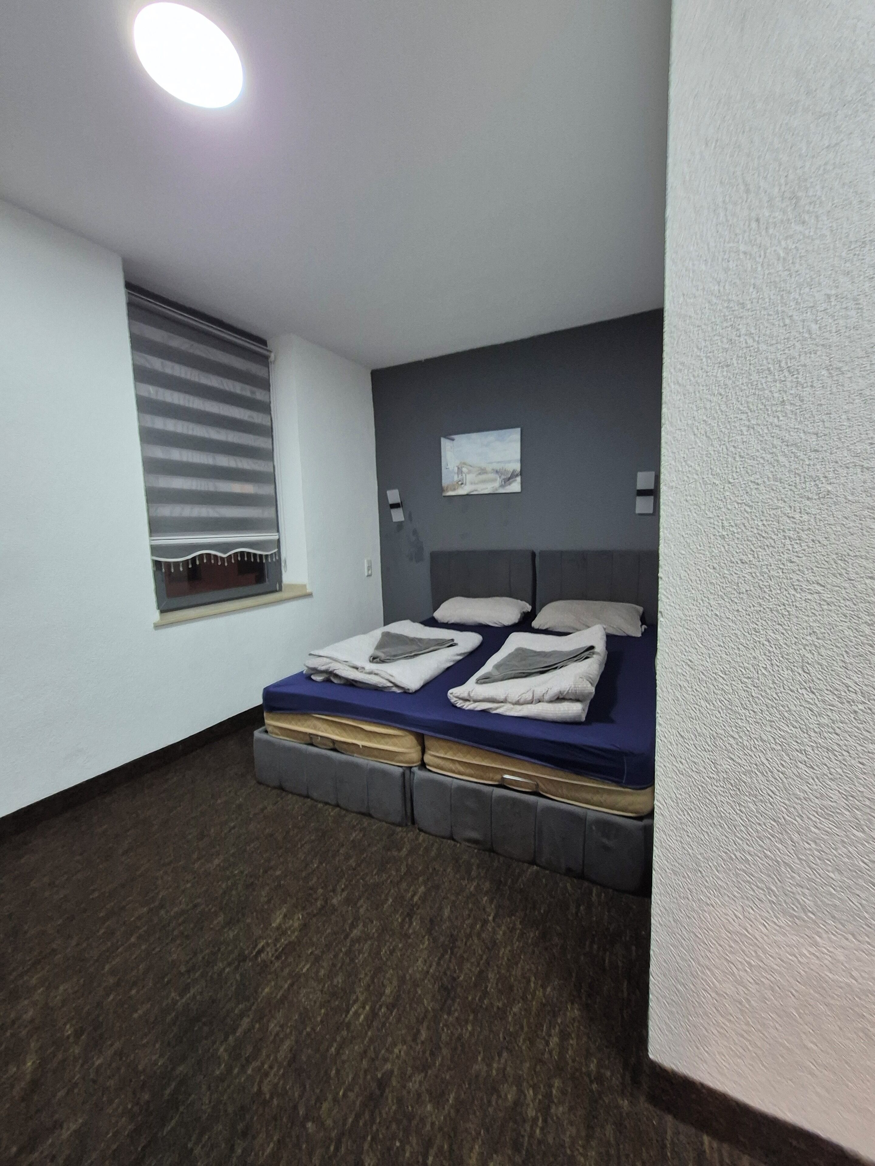 Ein Schlafzimmer mit Bett, einem Fenster mit Jalousien, einem kleinen Bild an der Wand und einer strukturierten Wand.