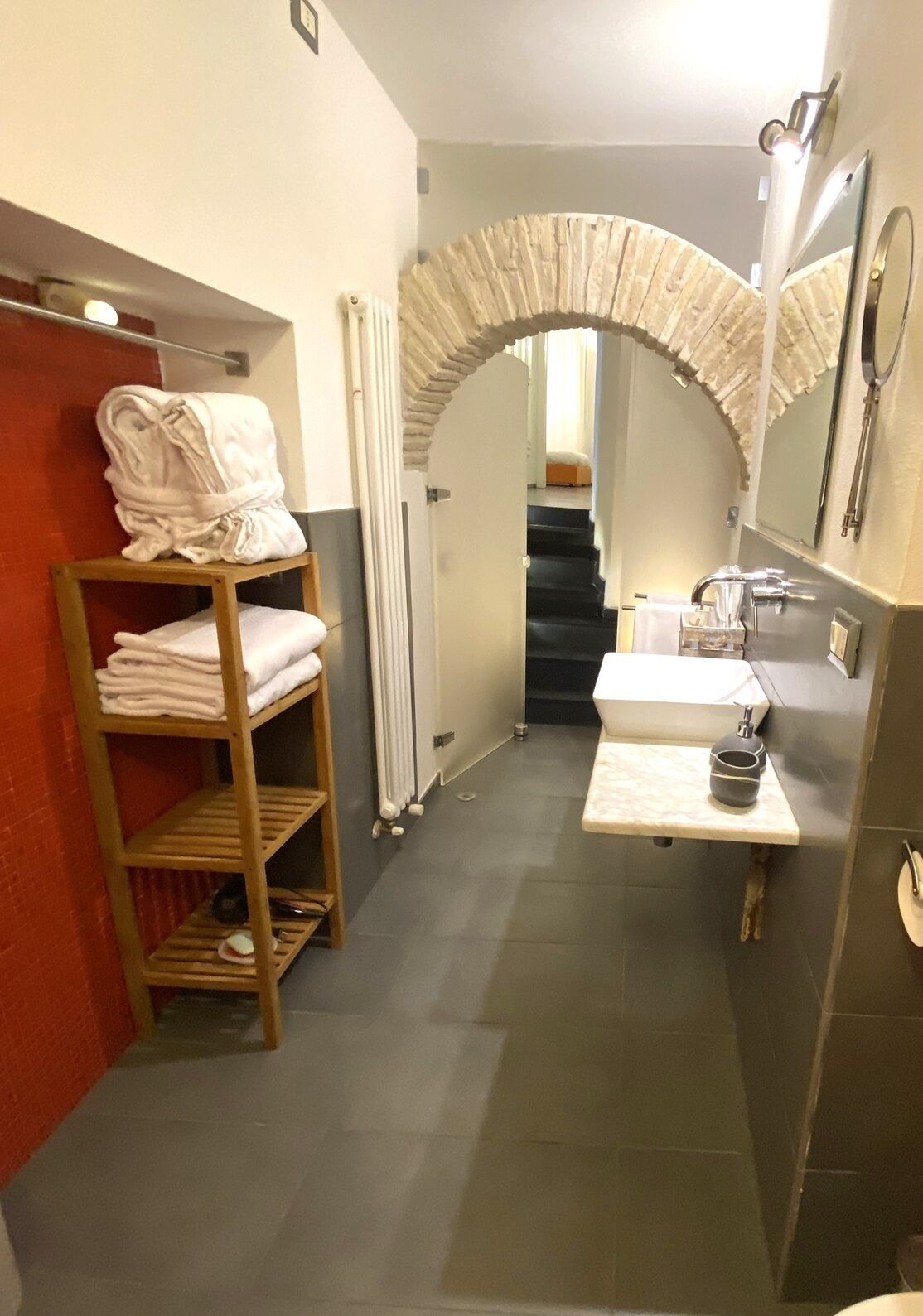 Un baño estrecho con un estante de madera que sostiene toallas, un lavamanos blanco con espejo y un arco de piedra que conduce a una escalera.