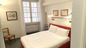 Un dormitorio con una cama roja, cortinas blancas, un ventanal con vistas, una silla y cuadros en la pared.