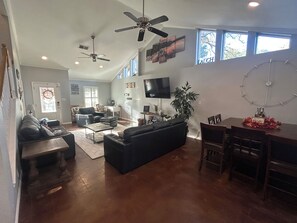 A spacious living area with a sofa, coffee table, and dining table. There are ceiling fans, a flat-screen TV, and a large window. - Huge property with Lake Access! 8 separate sleeping spaces, 4 baths! (Kingsland)