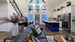 A spacious kitchen with a central island, a dining area with a black leather couch, and a staircase leading to an upper level. - HopSquatch: Air Hockey, Hot Tub, Games, Fire Pit, Pet-Friendly, Family-Oriented (Broken Bow)
