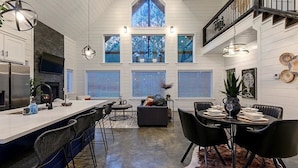 A modern kitchen with a central island, dining area with black chairs, and a staircase leading to an upper level. - HopSquatch: Air Hockey, Hot Tub, Games, Fire Pit, Pet-Friendly, Family-Oriented (Broken Bow)