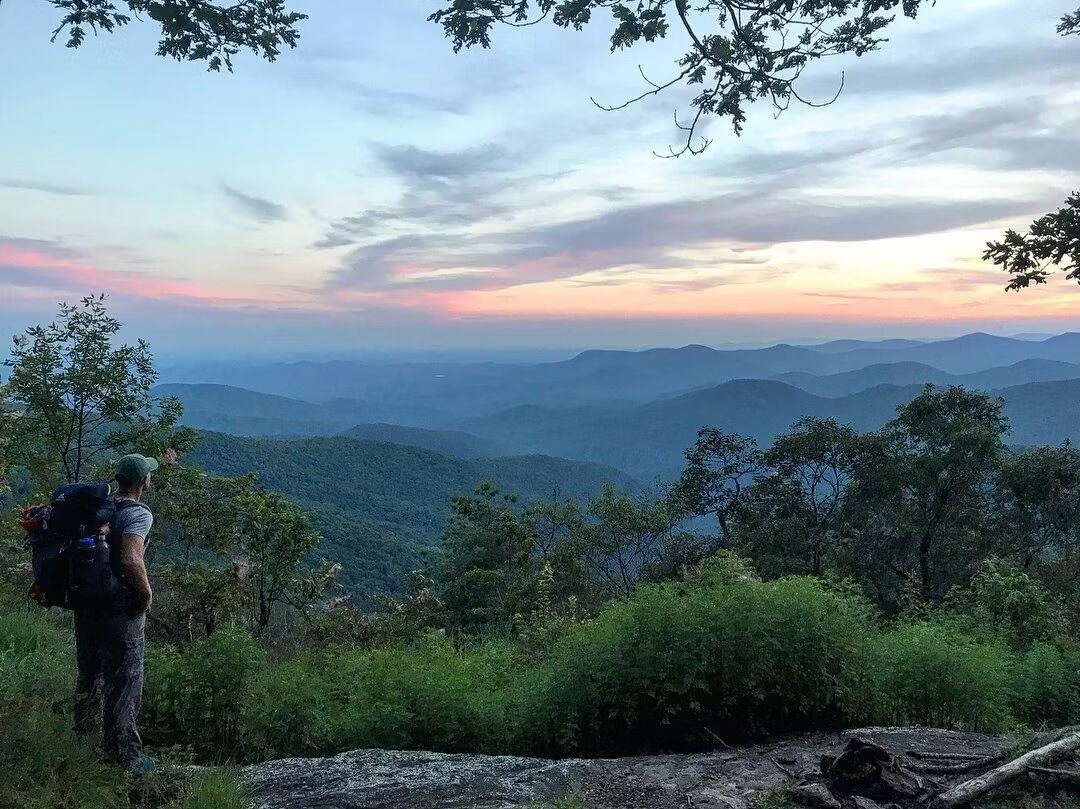 A hiker on a mountain trail with a view of distant mountains and a sunset.