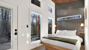 A bedroom with a large bed, white walls, and a view of trees through the windows. - Luxurious Modern Cabin - Timber For Two (Blue Ridge)