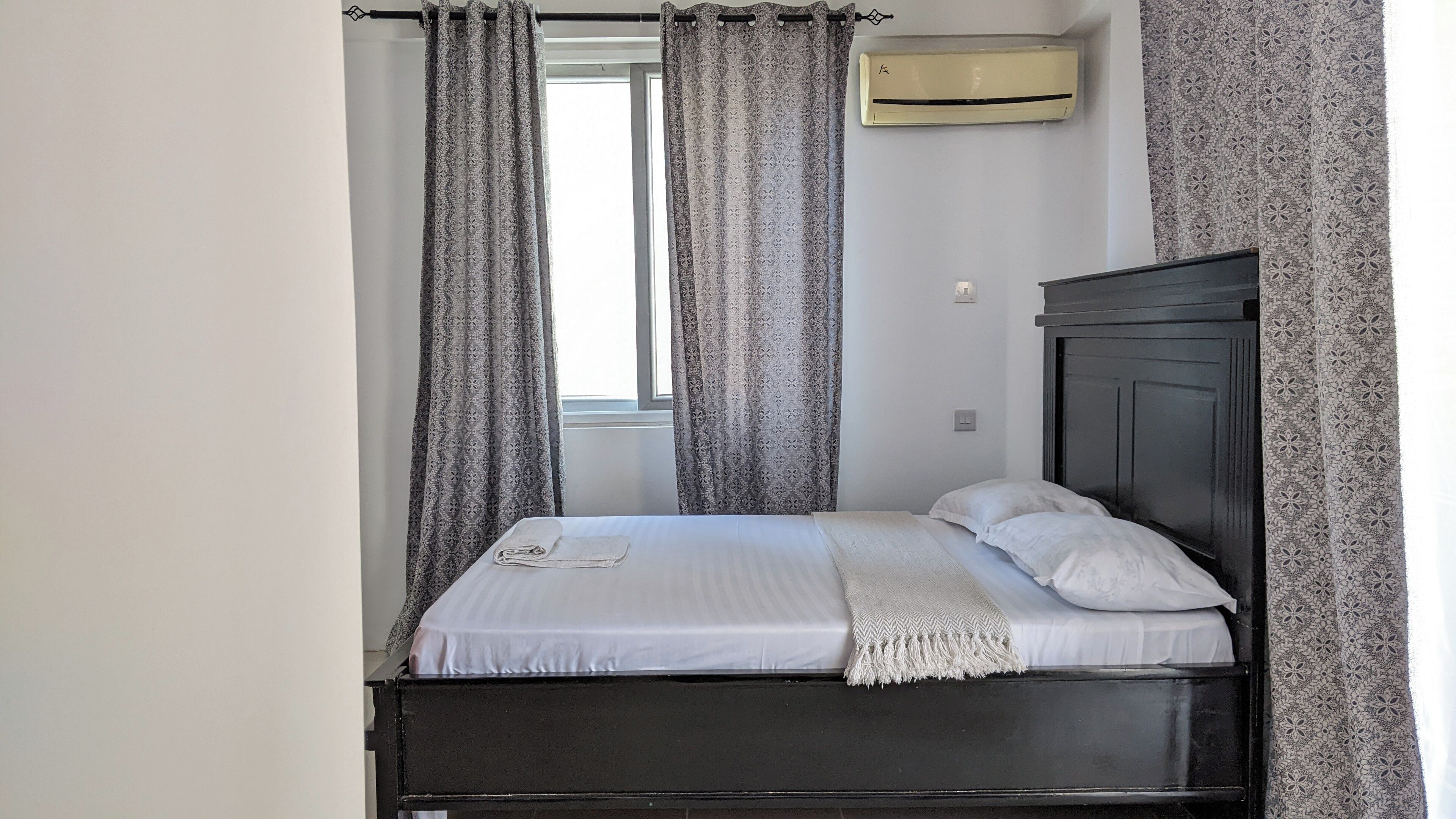 Un lit simple avec une tête de lit sombre, du linge de lit blanc et un plaid blanc. Il y a une fenêtre avec un rideau, et un appareil de climatisation fixé au mur.
