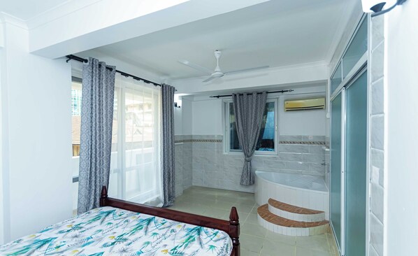 Un dormitorio con cama, ventilador de techo, ventana con cortinas, baño con bañera y retrete.