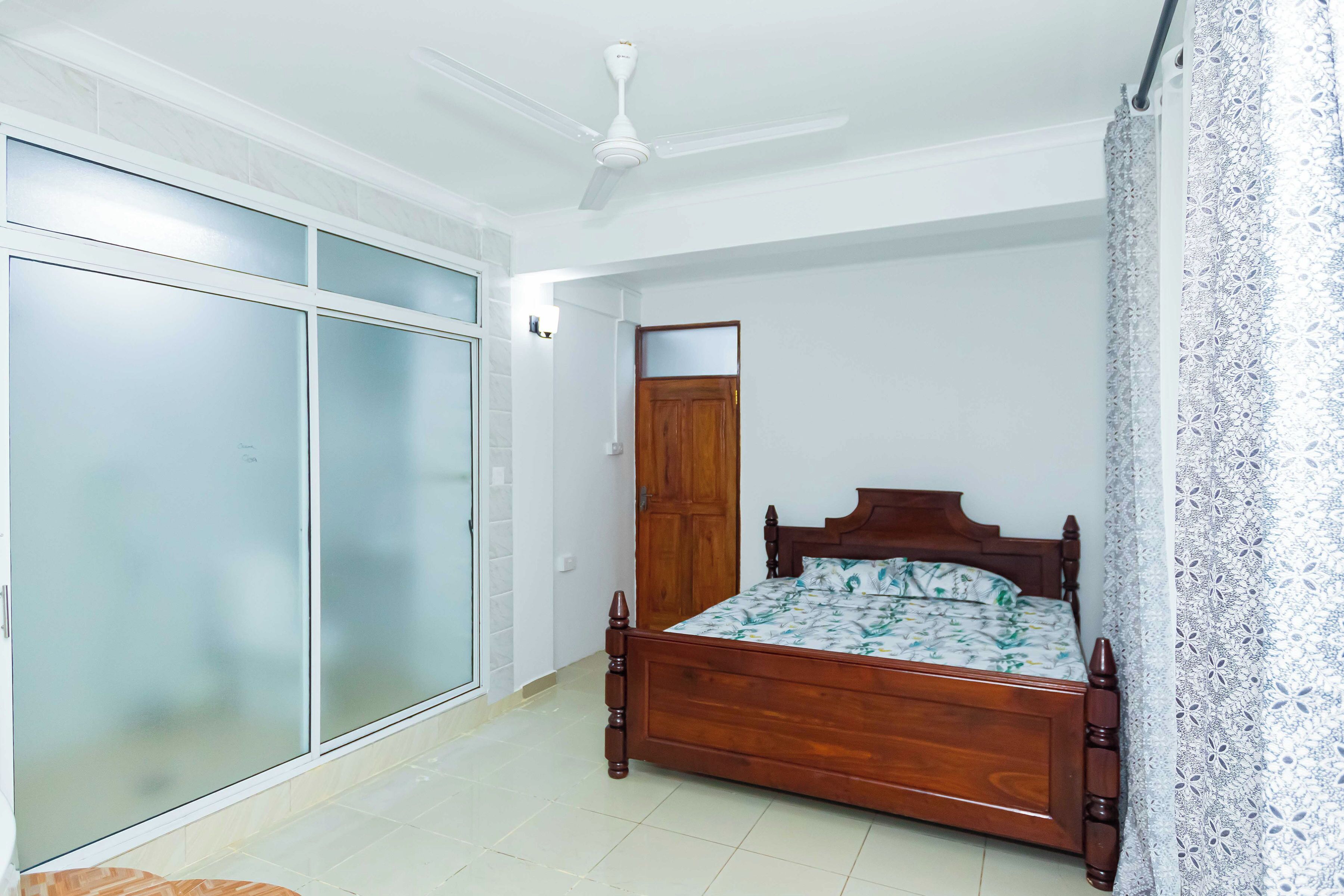 A bedroom with a wooden bed, a ceiling fan, a wardrobe with frosted glass doors, and patterned curtains.