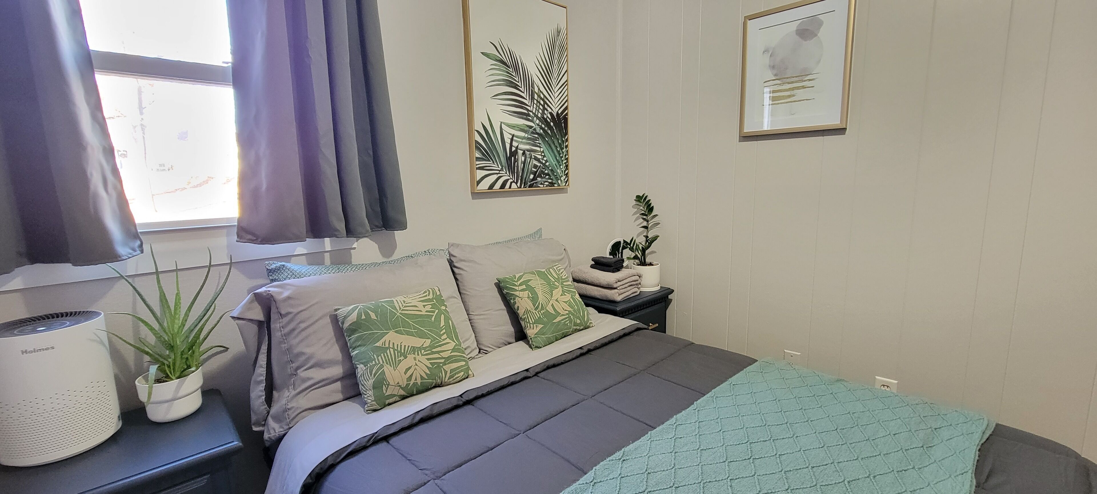 A bedroom with a bed, a bedside table with a plant, a heater, and a window with curtains.