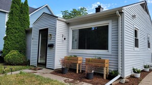 Una casa de dos pisos con un ventanal grande, un banco de madera y plantas en macetas.