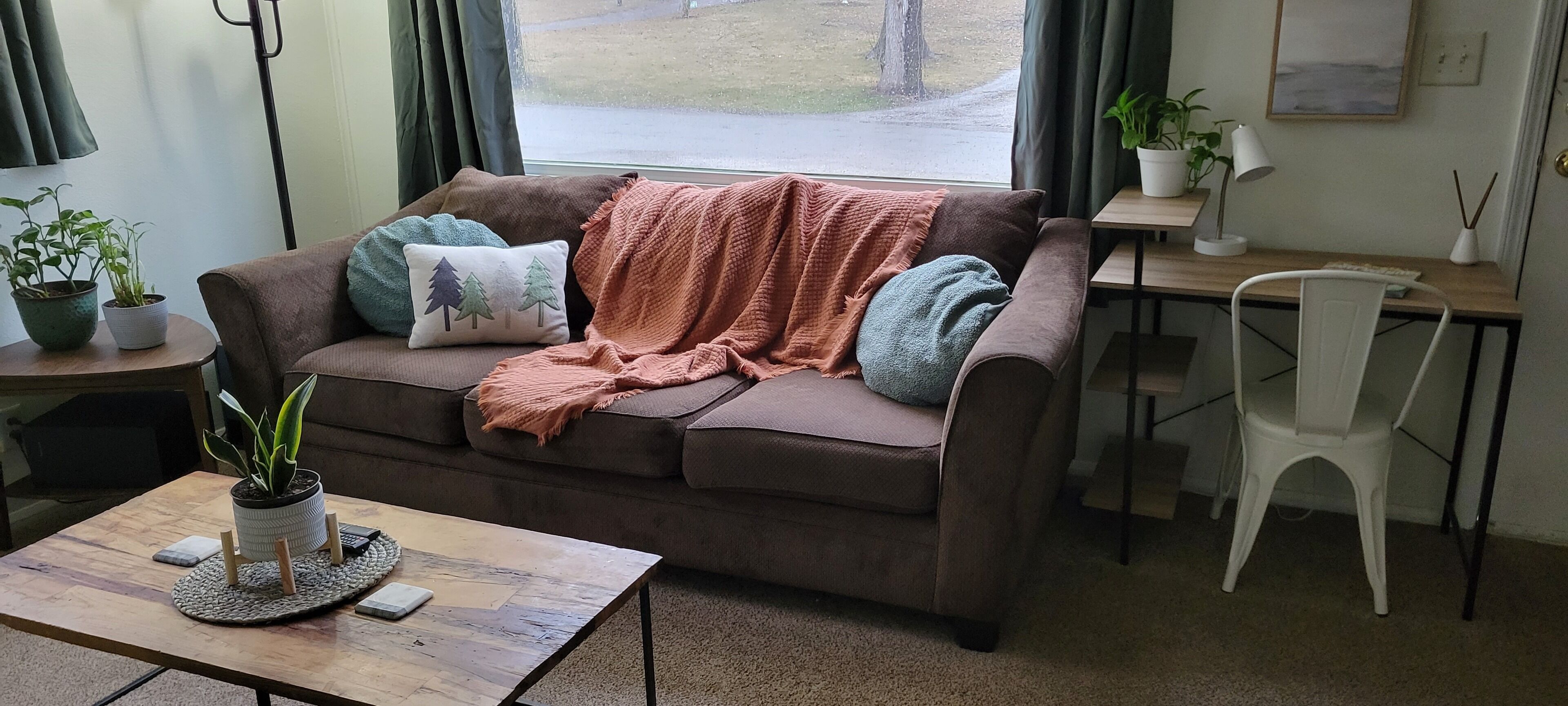 A cozy living room with a brown sofa, a coffee table, a desk, and a chair. There are potted plants on a side table and a window with a view of the outdoors.
