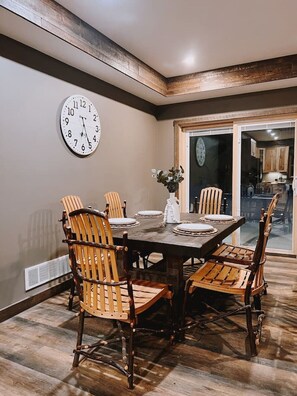 A dining area with a wooden table and chairs, a clock on the wall, and a view into the kitchen through a glass door. - Cozy two bedroom cottage with indoor fireplace. (Crosslake)