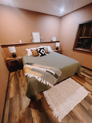 A bedroom with a bed, nightstands, a bedside table, a window, and a wooden floor. - Cozy two bedroom cottage with indoor fireplace. (Crosslake)