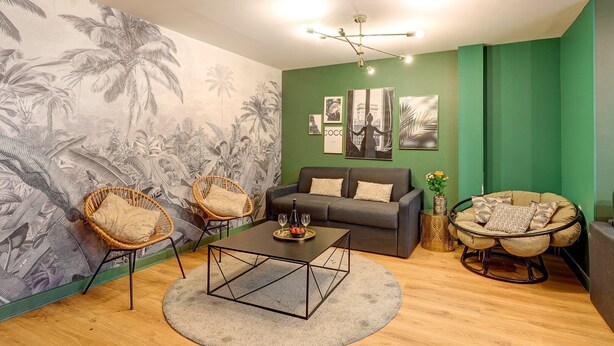 Ein Wohnzimmer mit grüner Akzentwand, einem großen tropischen Wandbild, einem grauen Sofa, zwei Korbstühlen, einem Couchtisch und einem Hängekorbstuhl.