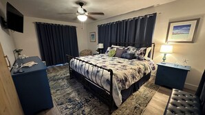 Ein Schlafzimmer mit einem Bett, einem Deckenventilator, einem Nachttisch, einer Lampe, einem gerahmten Bild und einem kleinen Tisch mit Dekoration.