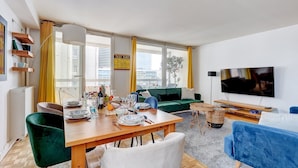 A modern living room with a dining area, large windows, a flat-screen TV, and a view of the cityscape. - 825 Suite Bacla - Superb apartment (Courbevoie)