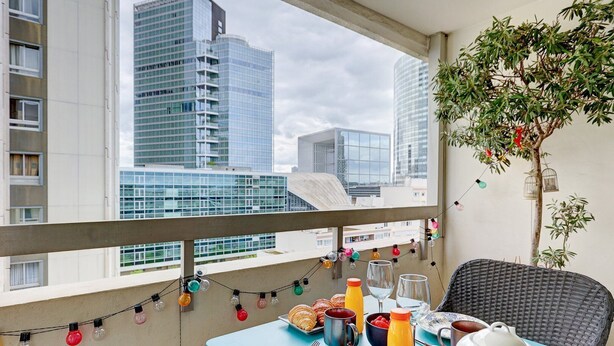 A balcony with a table set for breakfast, a potted plant, and a view of city buildings. - 825 Suite Bacla - Superb apartment (Courbevoie)