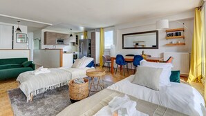 A modern, well-lit room with a bed, two sofas, a dining table with chairs, a kitchen area, and a large mirror. - 825 Suite Bacla - Superb apartment (Courbevoie)