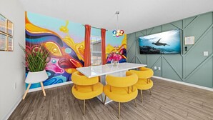 Ein moderner Essbereich mit einem weißen Tisch und gelben Stühlen, einem farbenfrohen Wandgemälde und einem an der Wand befestigten Fernseher.