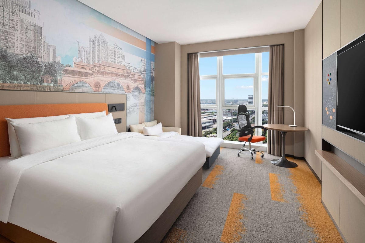 A hotel room with a large bed, a desk, a chair, a TV, and a window with a city view.
