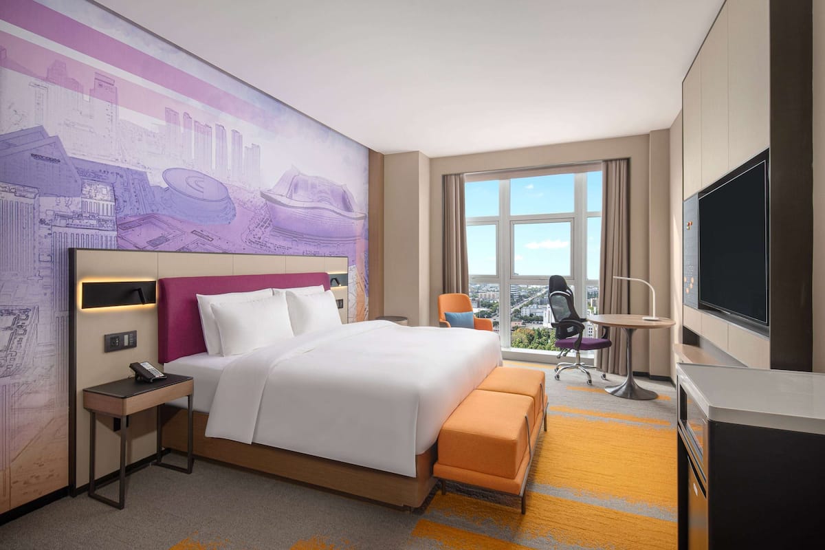 A hotel room with a large bed, a desk, a chair, a TV, and a window with a city view.