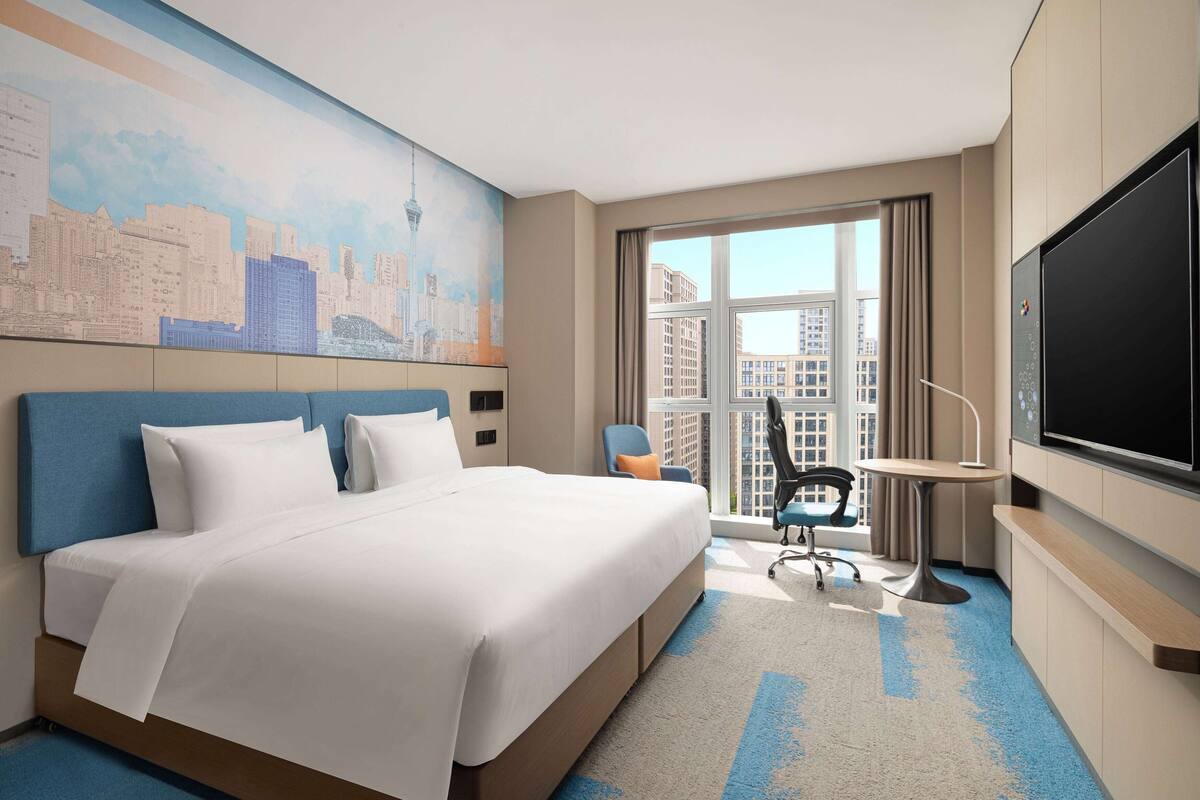 A hotel room with a large bed, a desk, a chair, a TV, and a window with a city view.