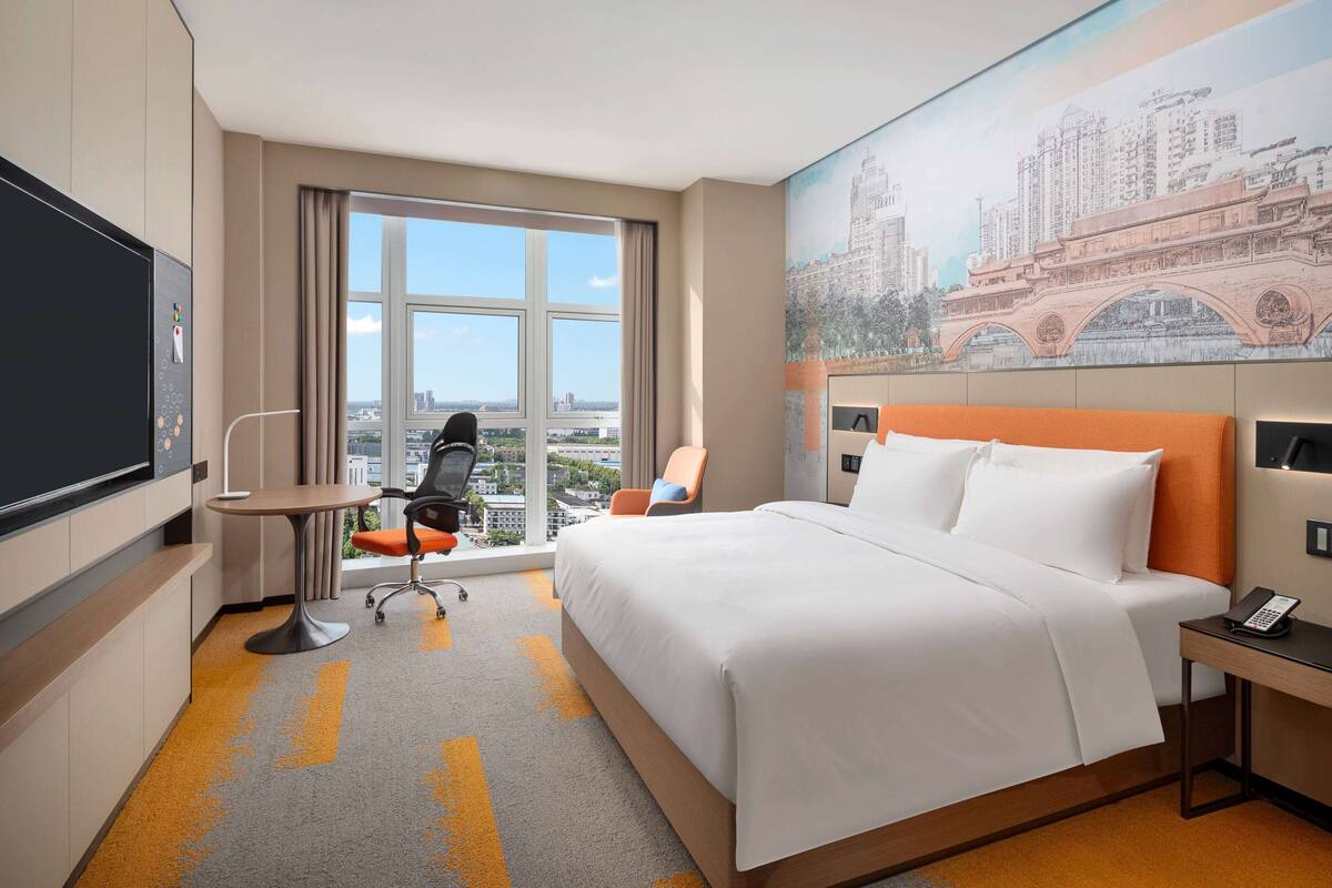 A hotel room with a large bed, a desk, a chair, a TV, and a window with a city view.