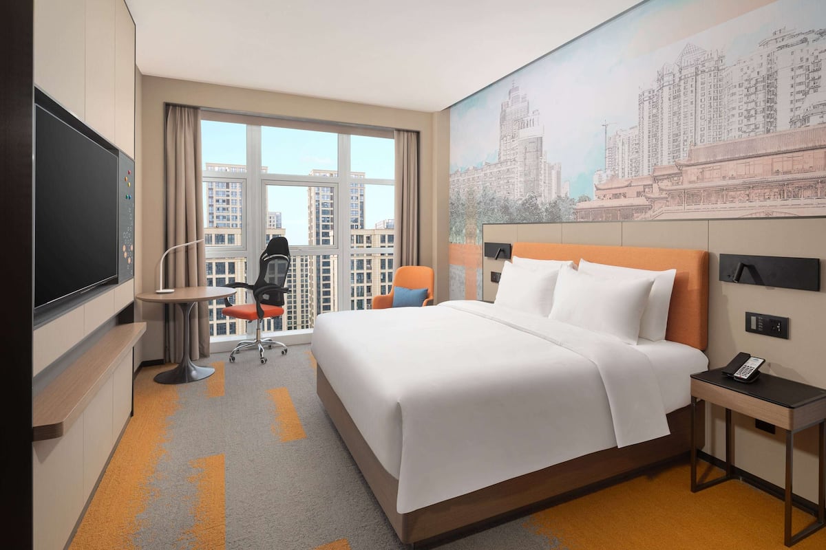 A hotel room with a large bed, a desk, a chair, a TV, and a window with a cityscape view.