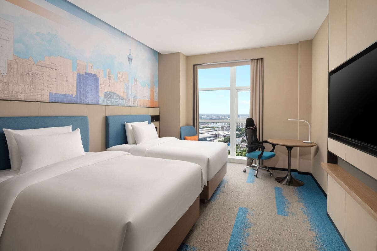 A hotel room with two beds, a desk, a chair, a large TV, and a cityscape mural on the wall.