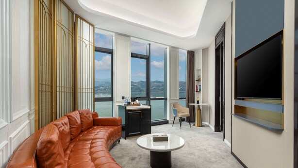 A modern hotel room with a leather sofa, a round coffee table, a desk, and a large TV. - Ramada By Wyndham Yichang Riverside (Yichang)