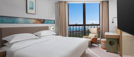 A hotel room with a large bed, a chair, a desk, a view of the city, and a painting on the wall.