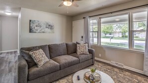 Un salon avec un canapé gris, deux coussins à motifs, un ventilateur de plafond, un tableau accroché au mur et une grande fenêtre donnant sur un quartier résidentiel.