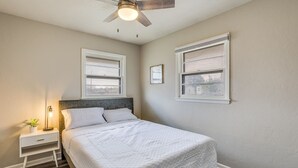 Une chambre à coucher comprenant un lit, une table de chevet, une lampe, un ventilateur de plafond et deux fenêtres.
