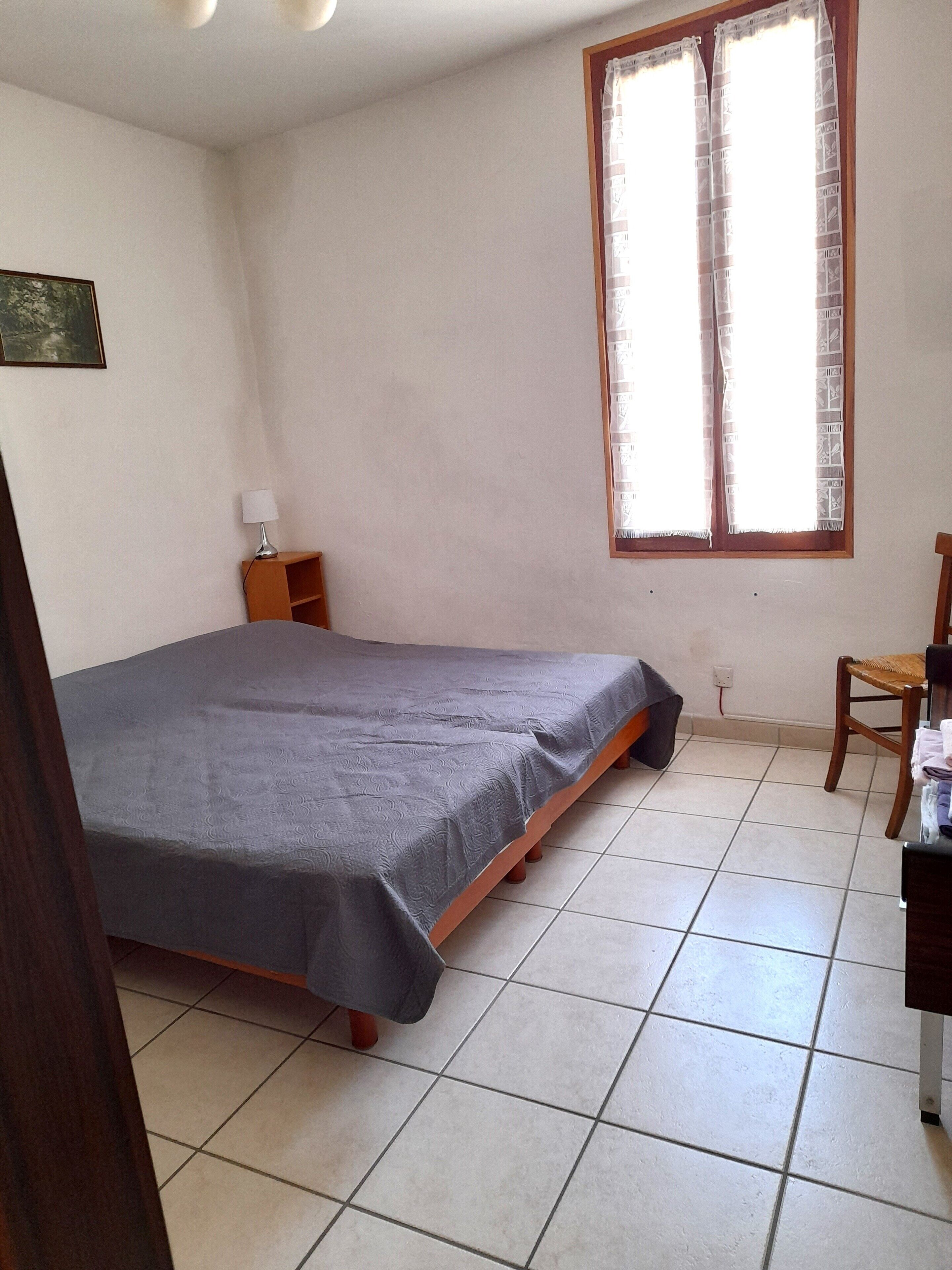 A small, tidy bedroom with a single bed, a nightstand, a chair, and a window with curtains.
