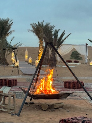 A desert campfire with a tent and palm trees in the background.