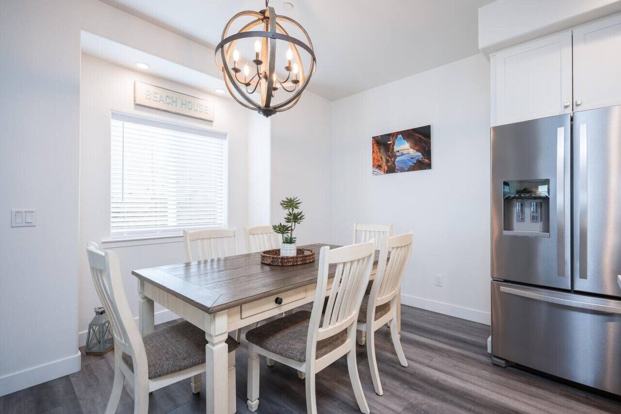 A dining area with a wooden table and white chairs, a chandelier, a refrigerator, and a window with blinds.