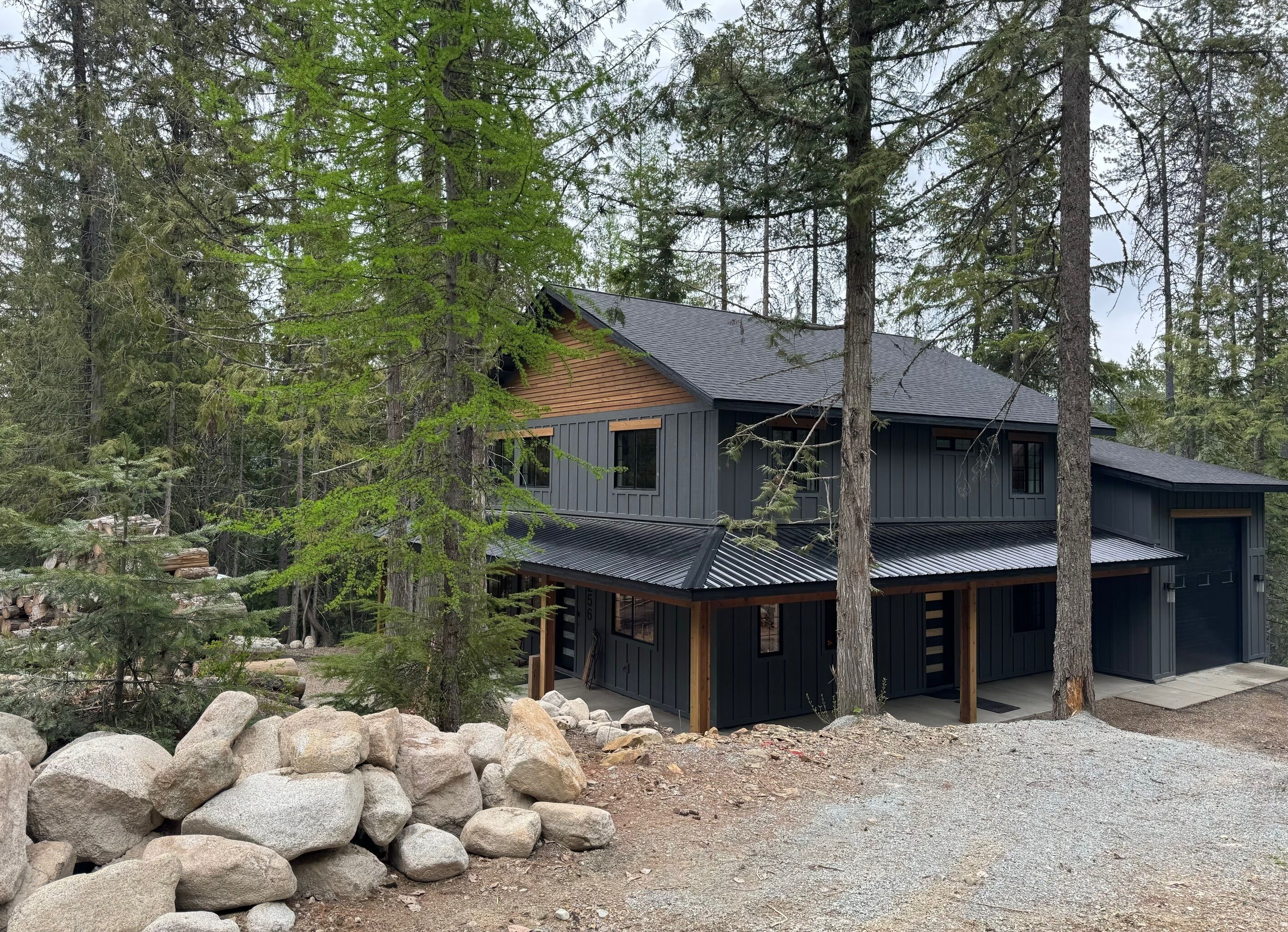 Una casa moderna de dos pisos con fachada oscura, rodeada de un bosque denso y grandes rocas.