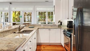 Una cocina moderna con armarios blancos, un refrigerador de acero inoxidable, un horno empotrado y un amplio ventanal con vistas a árboles.