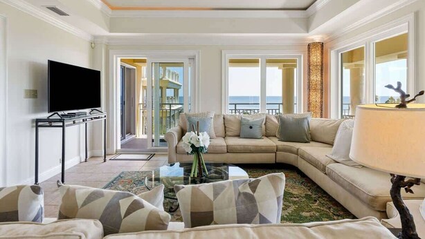 A spacious living room with a large sofa, a coffee table, and a TV. A balcony with a view of the sea is visible through an open door.