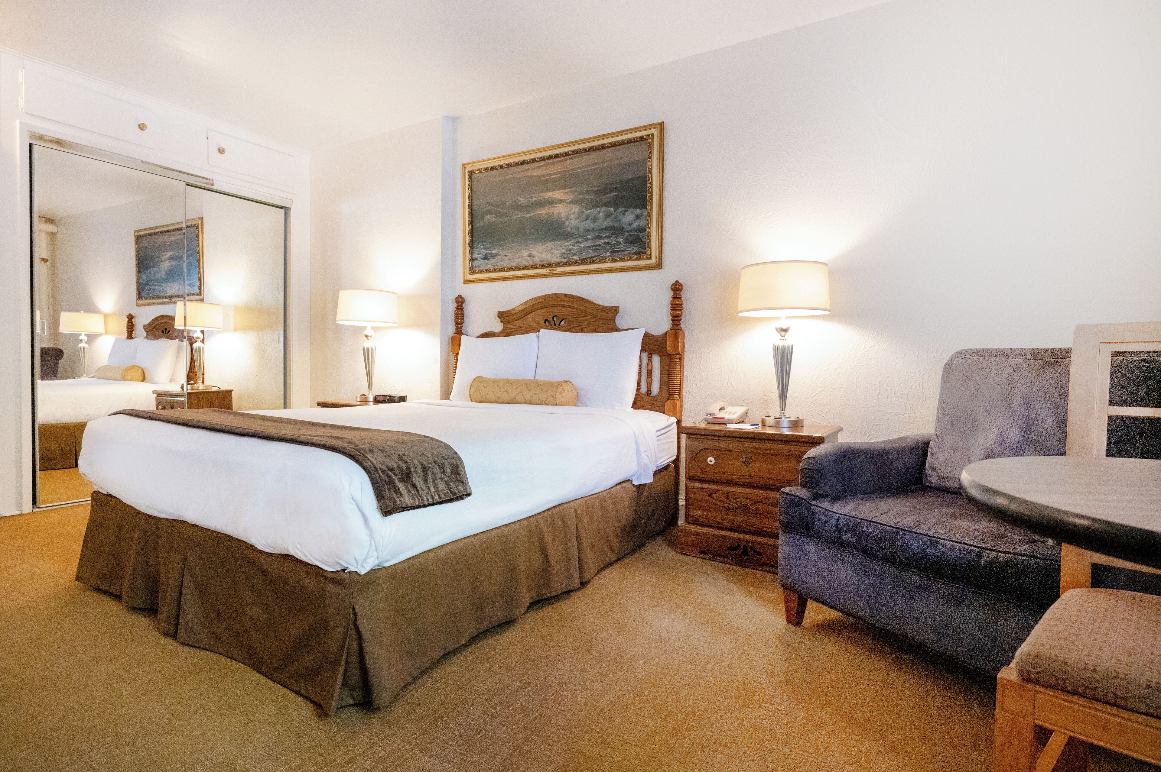 A hotel room with a large bed, a wooden nightstand with a lamp, a chair, a small table, and a painting on the wall.