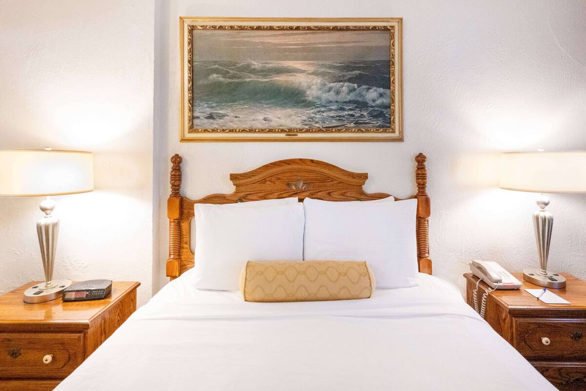 A neatly made bed with a wooden headboard, two bedside tables with lamps, a framed picture of the sea, and a small bedside table with a phone and a remote control.