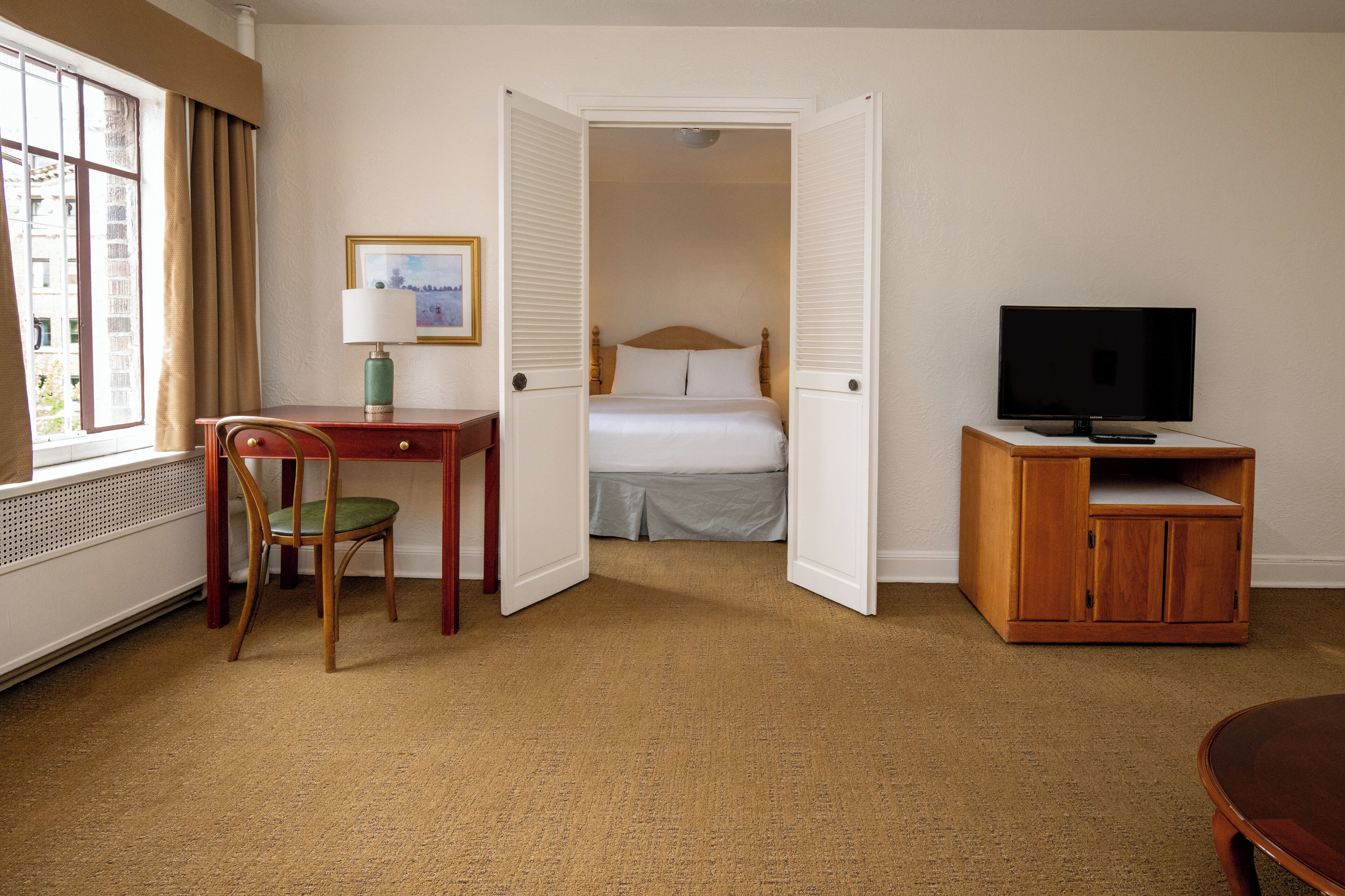 A hotel room with a bed, a desk with a chair, a television on a wooden stand, and a window with curtains.