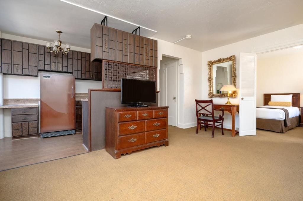 A compact hotel room with a kitchenette, a wooden dresser, a desk with a chair, a bed, and a large mirror.