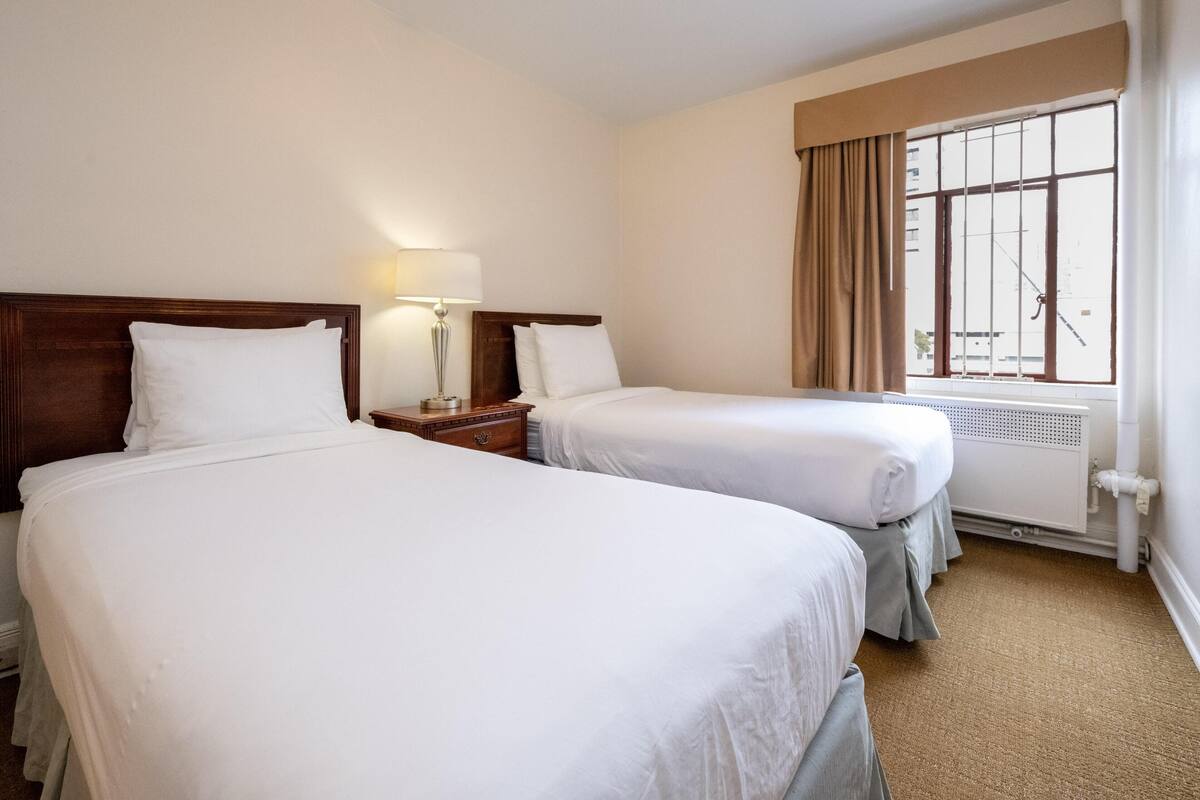 A hotel room with two single beds, a nightstand with a lamp, a window with curtains, and a radiator.