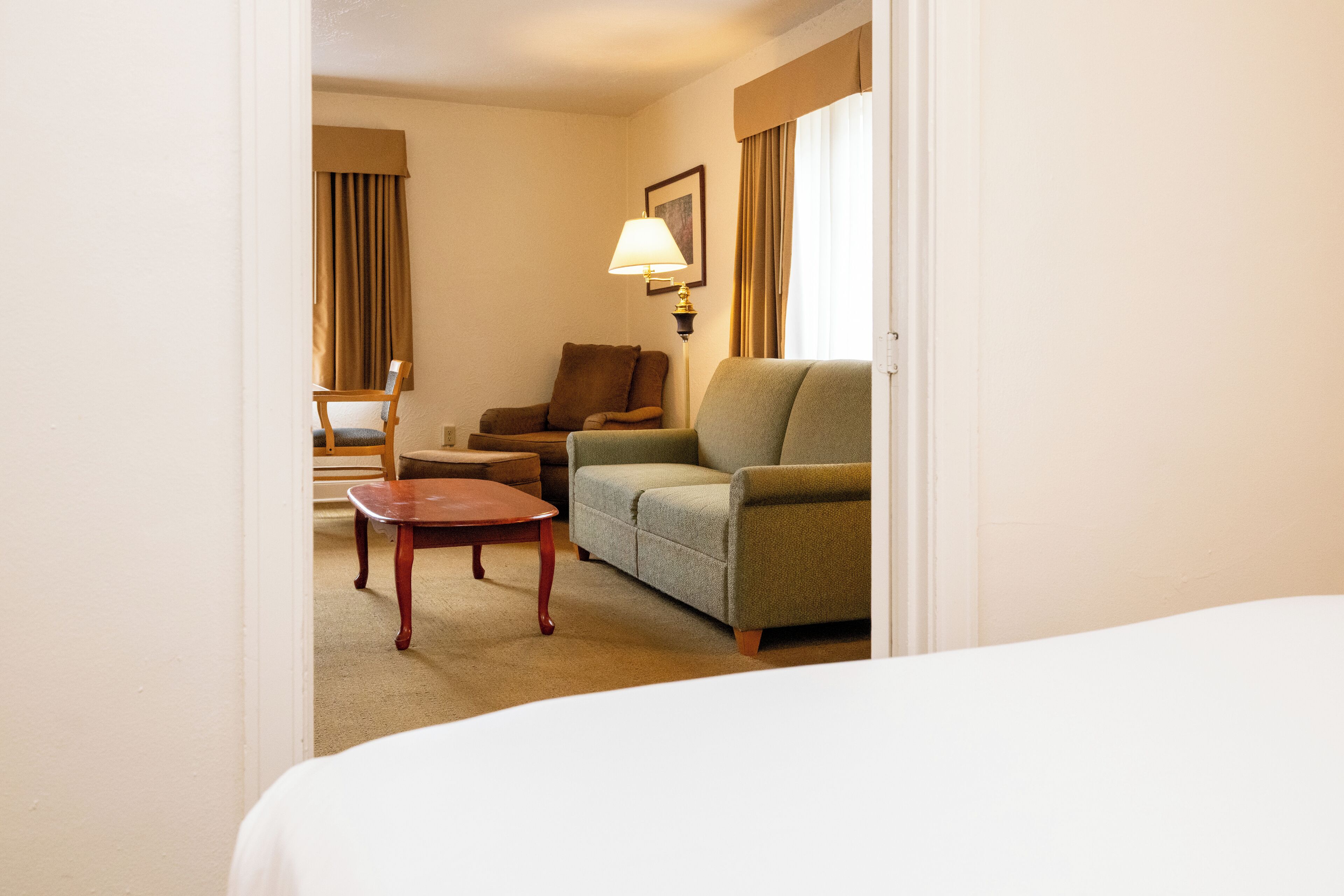 A hotel room with a bed, a green sofa, a wooden chair, a small table, and a lamp.