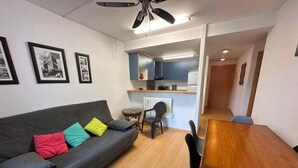 A compact living space with a grey sofa, colorful cushions, a wooden dining table, and a small kitchen area. - Apartment with communal swimming pool AV 29 (Rosas / Roses)