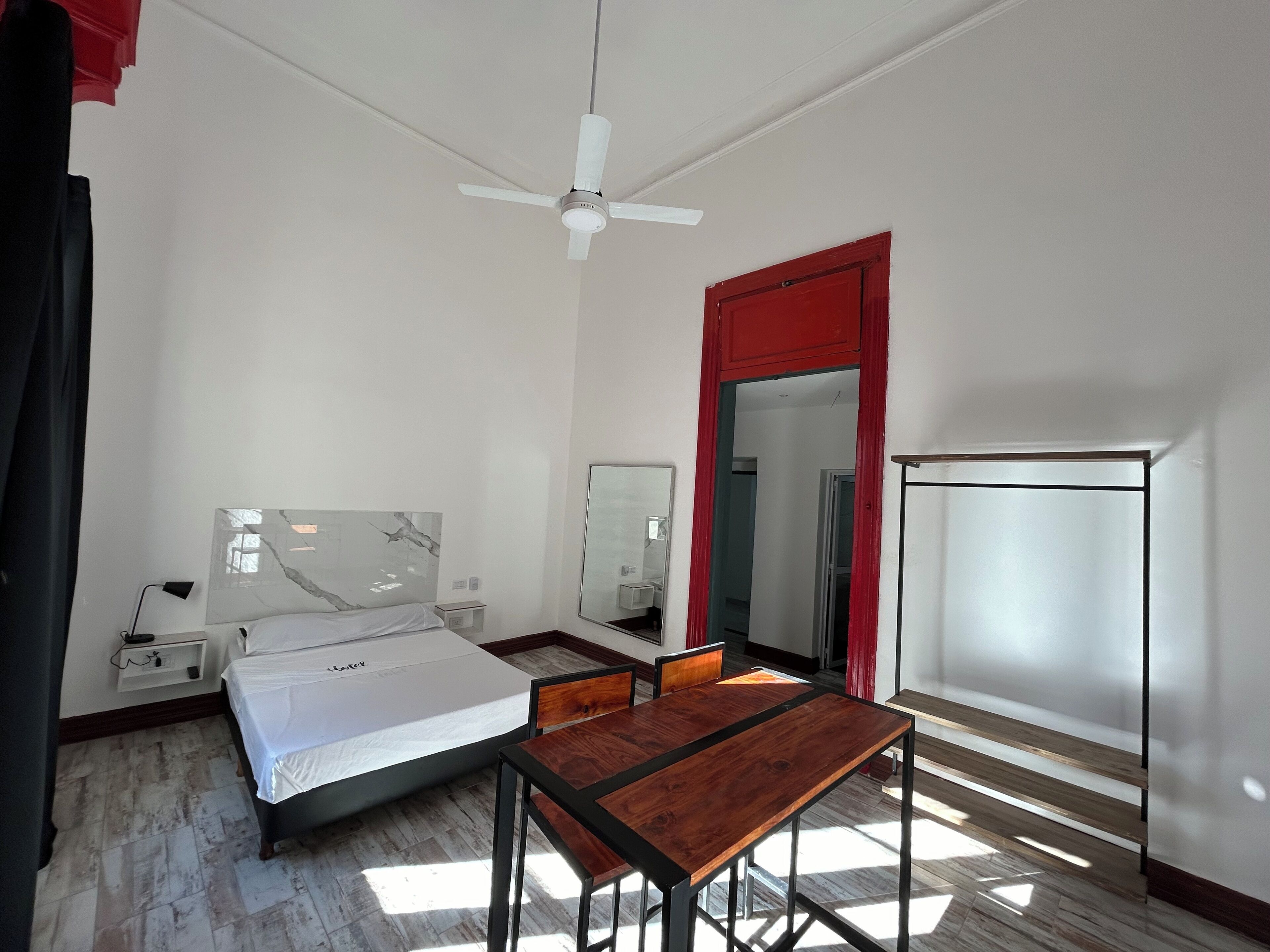 A hotel room with a bed, a desk, a chair, a mirror, a ceiling fan, and a refrigerator.