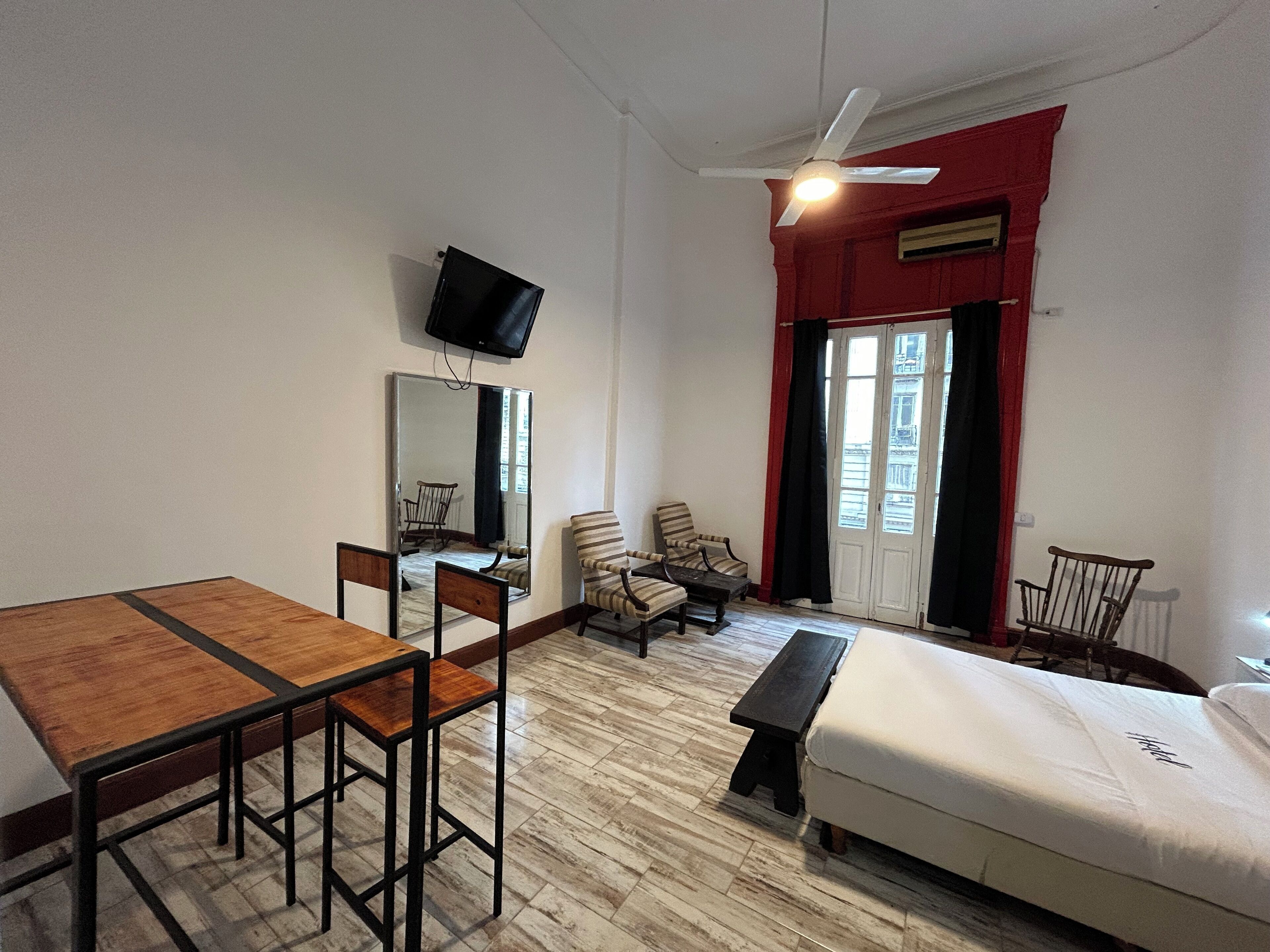 Ein Hotelzimmer mit einem Holztisch, vier Stühlen, einem Einzelbett, einer Bank, einem an der Wand befestigten Fernseher und einem Fenster mit roten Vorhängen.