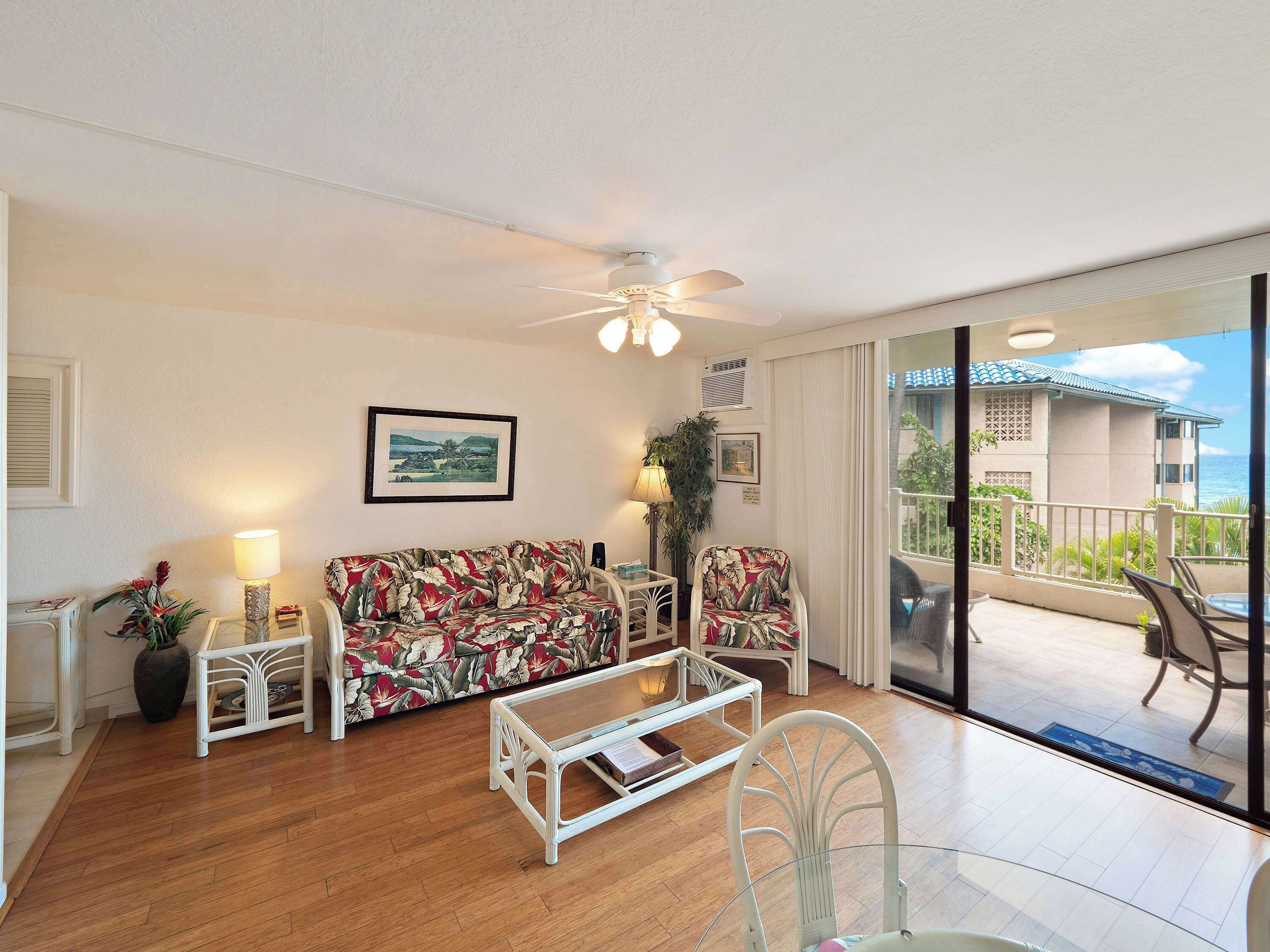 A living room with a sofa, chairs, a coffee table, and a ceiling fan. There is a view of a balcony with outdoor furniture.